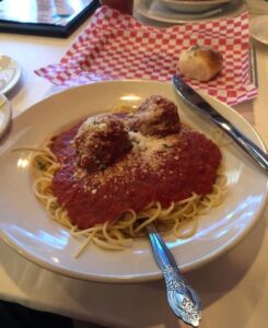 Spaghetti with red sauce, 2 meatballs and parm cheese on a white plate with red and white checkered napkin and bread roll behind it